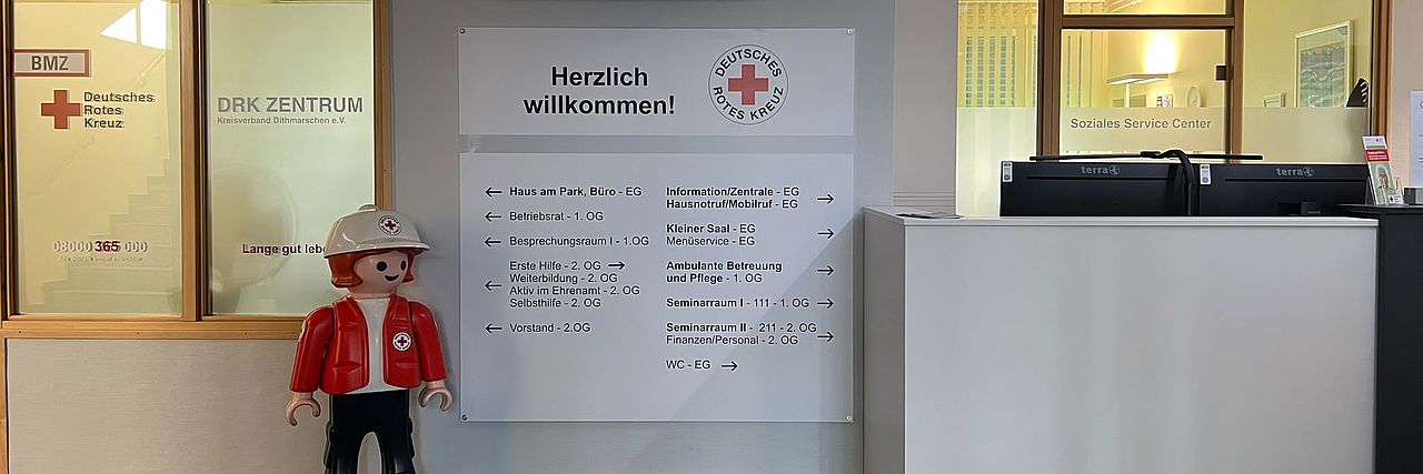 Blick in den Eingangs-/Tresenbereich. Links Soziales Service Center. Kleine Playmobilfigur, Kind in DRK Kleidung. Helle Übersichtstafel mit Hinweisen zu den Bereichen im Kreisverband. Rechts Teile des Empfangstresen